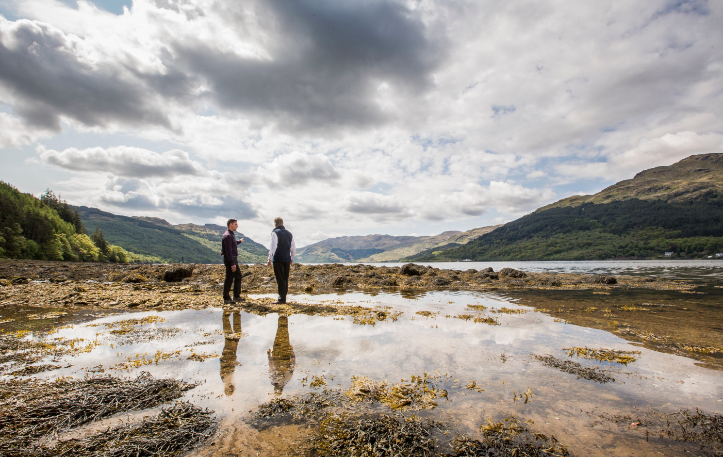 Two people stand on reflective wet rocks overlooking a scenic mountain lake under dramatic cloudy skies in Scotland.