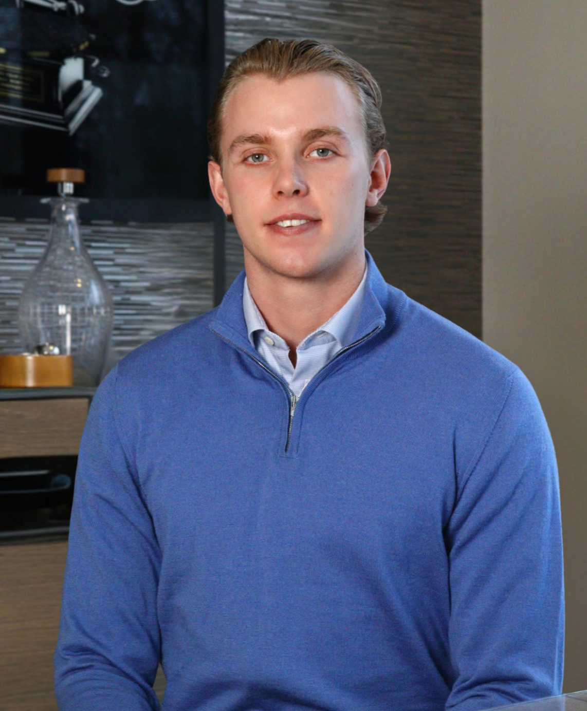 Young professional man smiling in blue quarter-zip sweater, posed indoors against modern interior backdrop.