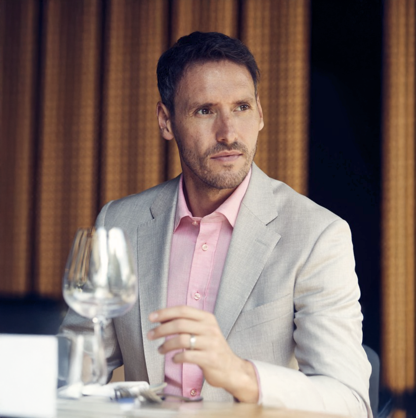 Man in pink shirt and gray blazer sitting at a white table with wine glasses in a modern restaurant setting.
