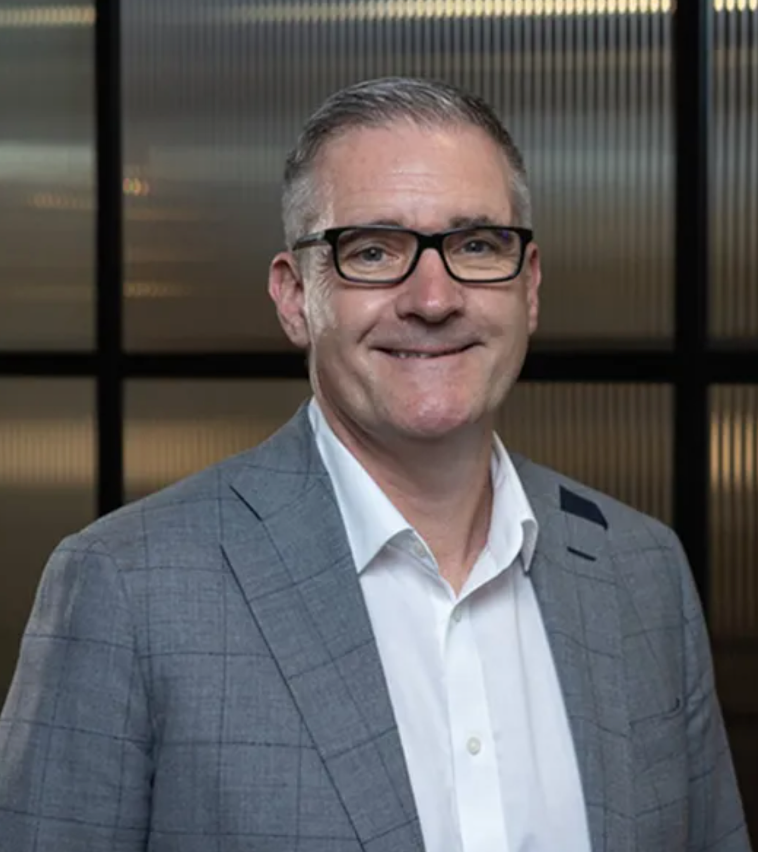 Professional businessman in gray blazer and glasses smiling confidently in a modern office setting.