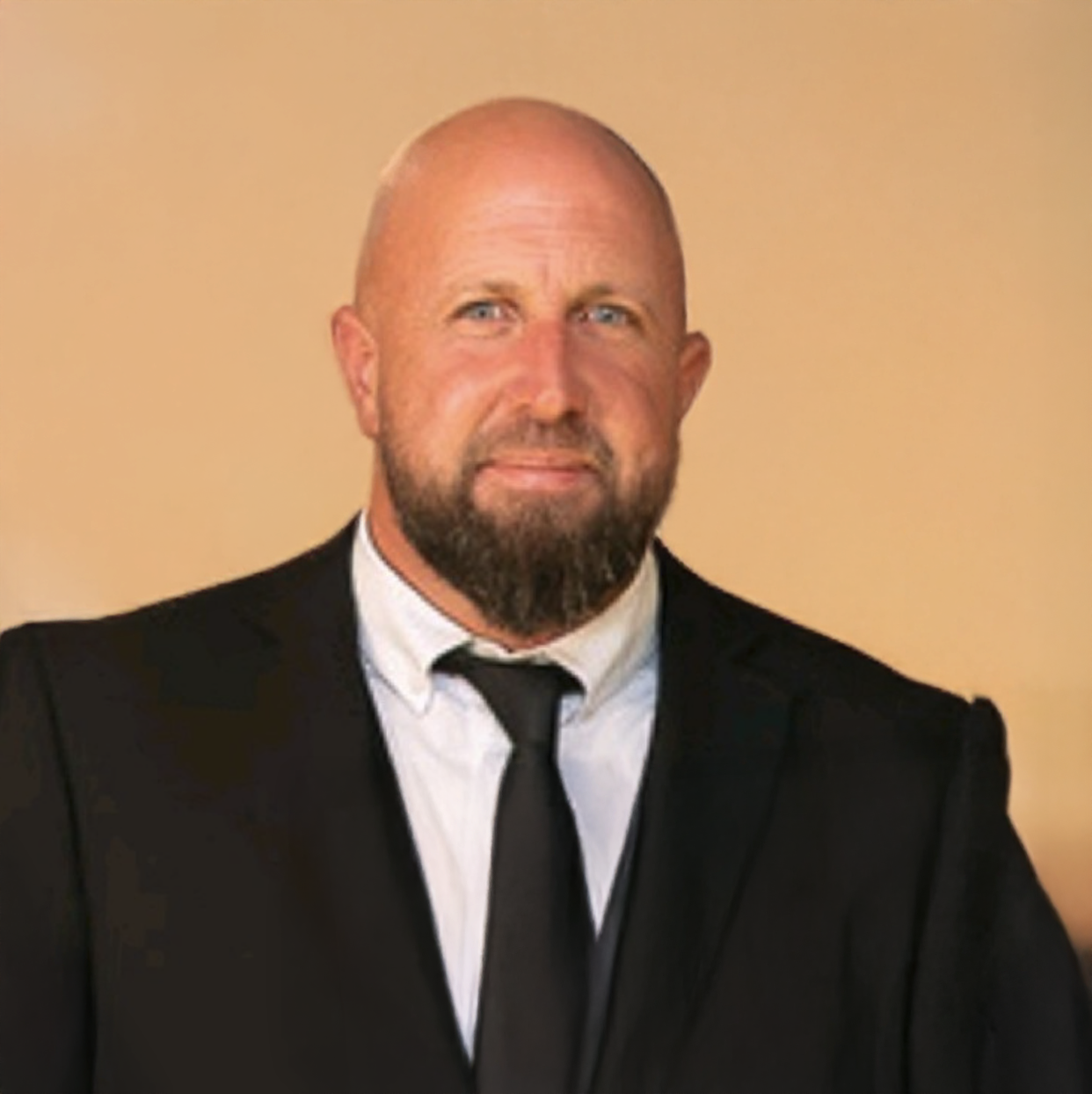 Bald man with a beard wearing a black suit and tie, posing professionally against a beige background.