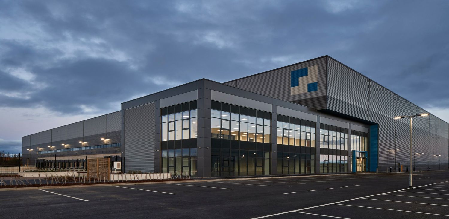 Modern industrial warehouse building with large windows and blue logo, photographed at dusk in an empty parking lot.