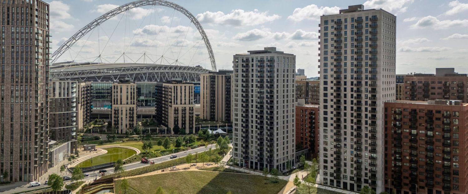Aerial view of Wembley Stadium arch rising above modern residential tower blocks and green spaces in London.