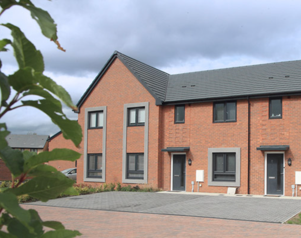Row of modern brick townhouses with grey roofs and white trim on a residential street with green trees in the foreground.