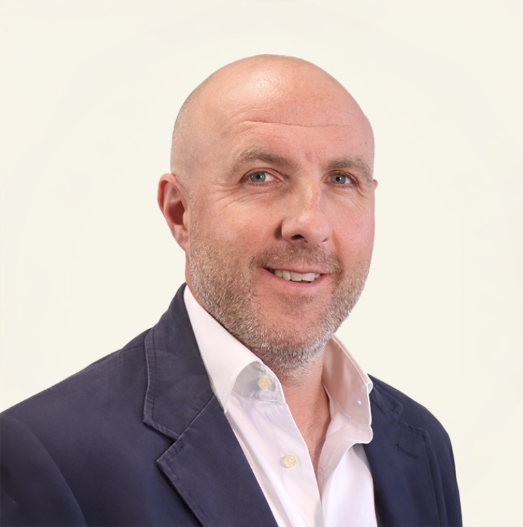 Smiling bald man with salt-and-pepper beard wearing a navy blazer and white shirt against a light background.