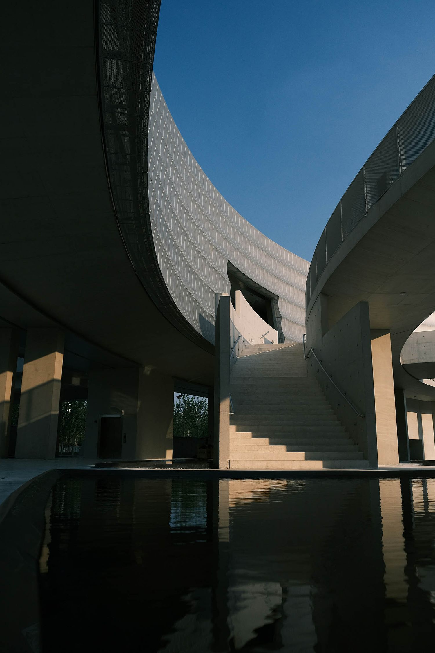 Modern curved concrete building with reflective pool and staircase under clear blue sky.