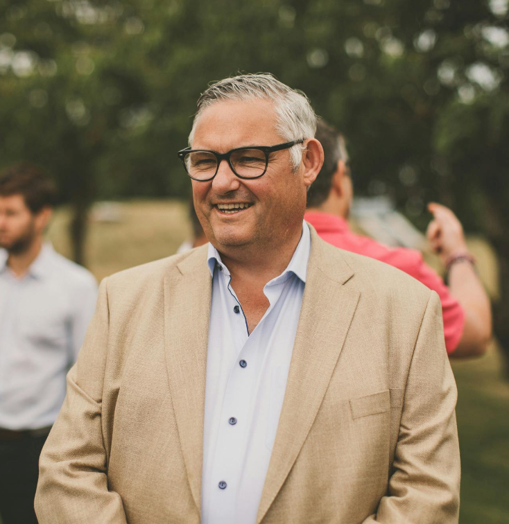 Smiling middle-aged man with glasses in a tan blazer outdoors at a social gathering.