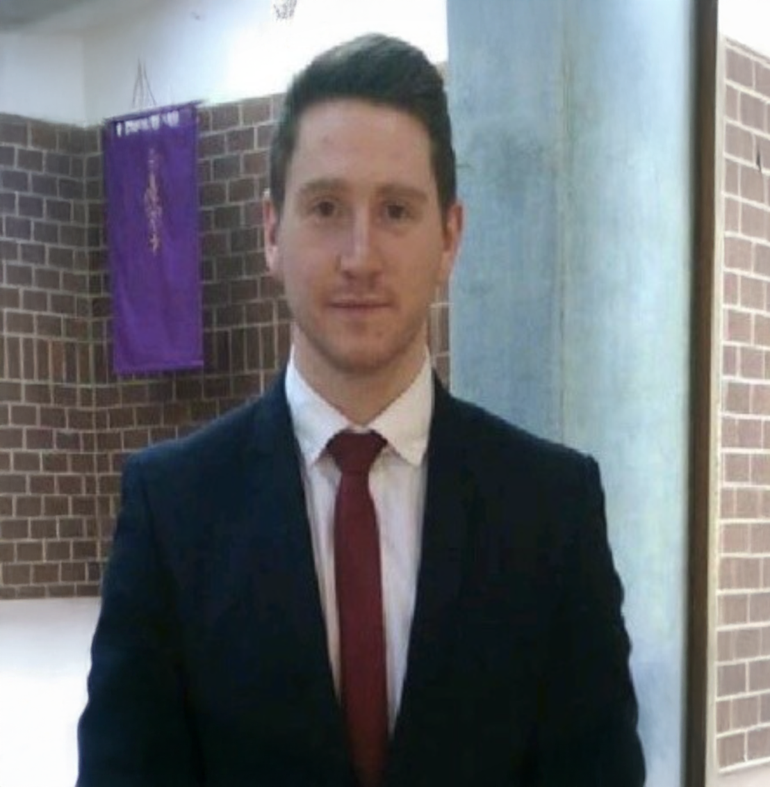 Professional man in dark suit and red tie posing indoors near brick wall with purple banner.
