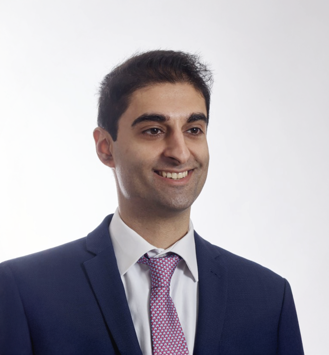 Young professional man in navy suit and pink tie smiling confidently against light gray background.