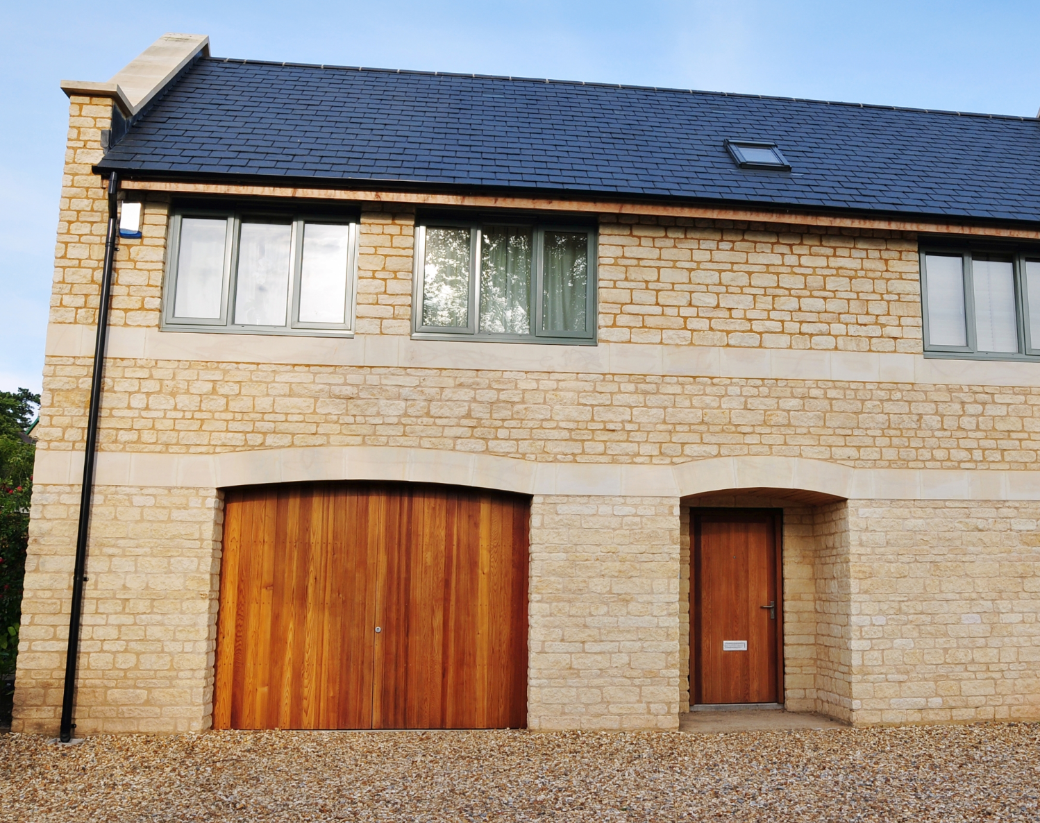 Modern stone house with wooden garage door and slate roof in Cotswold style architecture.