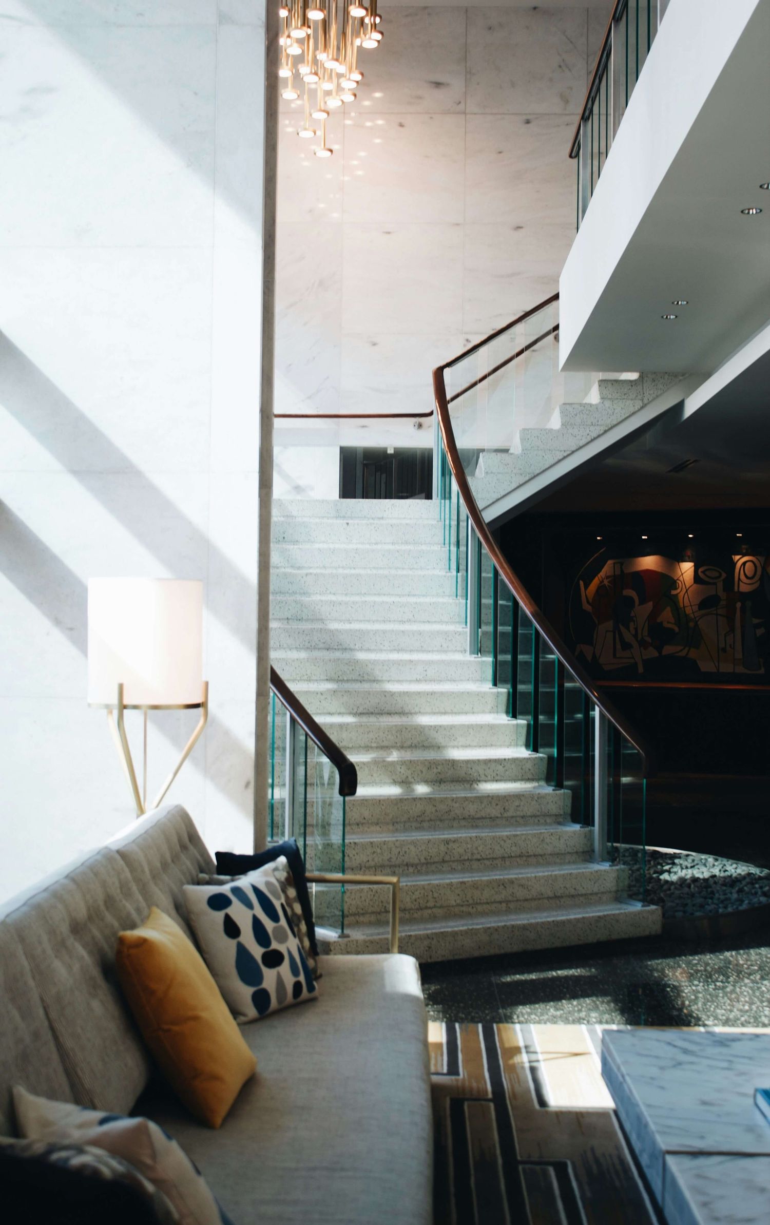 Modern hotel lobby with marble staircase, glass railings, pendant lights, and a cozy sofa with decorative pillows.
