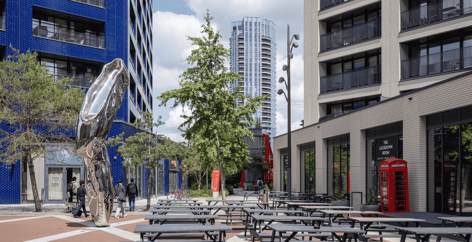 Modern urban plaza with picnic tables, red phone box, sculpture, and city skyscrapers on a sunny day.