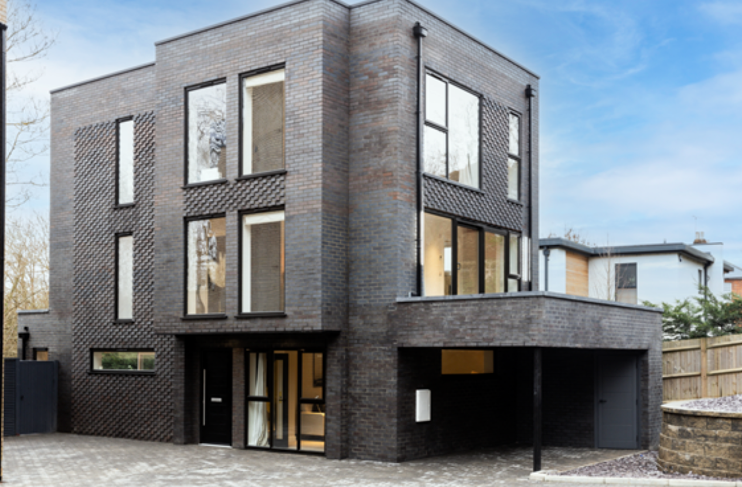 Modern three-story dark brick house with large windows, carport, and contemporary architecture in a residential setting.
