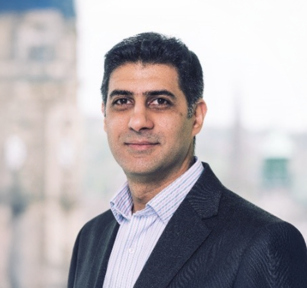 Professional headshot of a middle-aged man in a dark blazer smiling outdoors with a blurred city background.