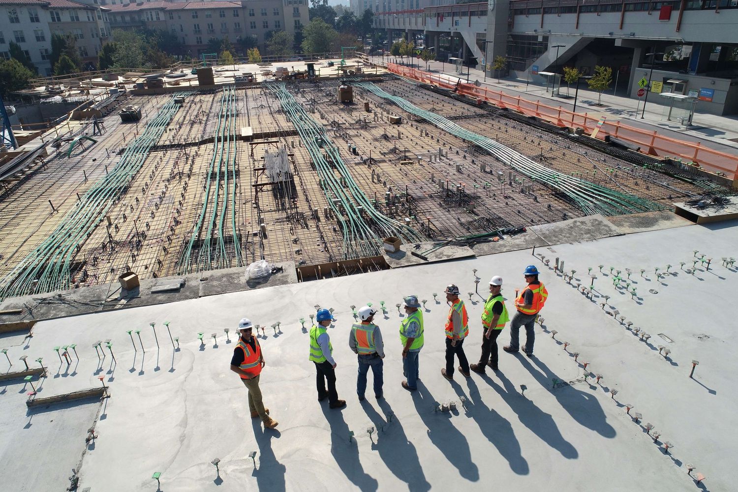 Construction workers in safety vests oversee a large building foundation site with steel reinforcement bars.