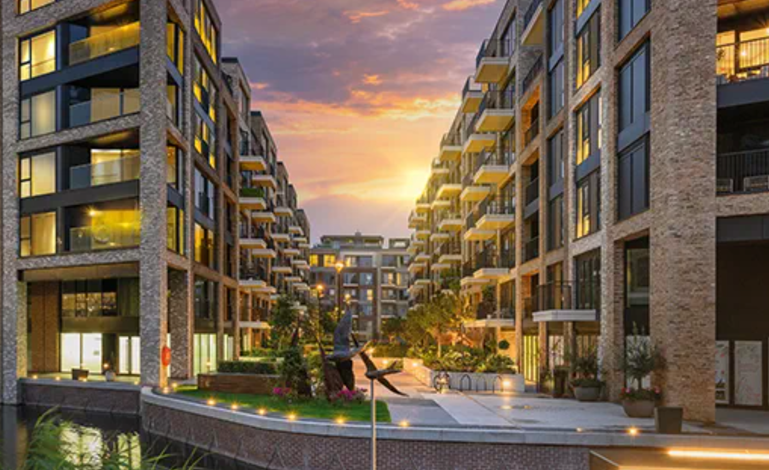 Modern luxury apartment complex with brick facades, balconies, and a serene courtyard water feature at sunset.