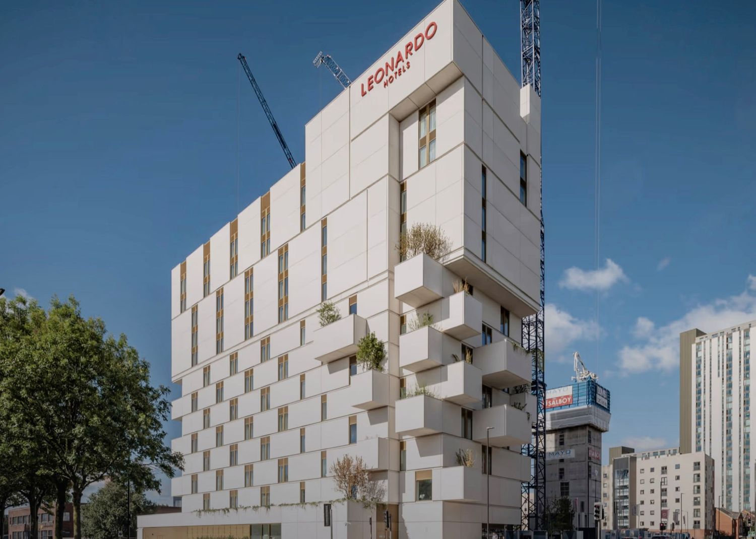 Modern Leonardo Hotel building with white geometric facade, balconies, and cranes against a blue sky in an urban setting.