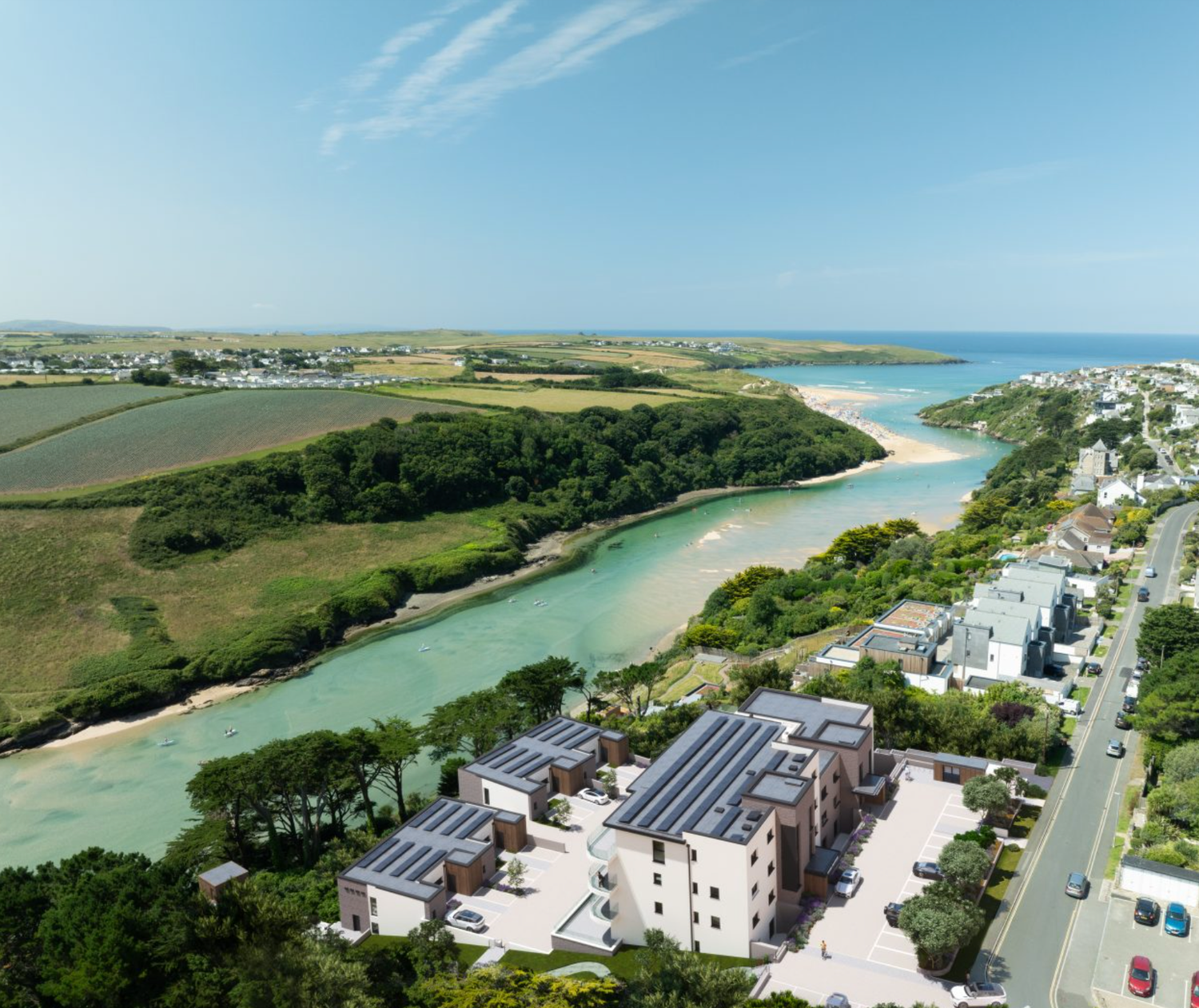 Aerial view of a coastal estuary with sandy shores, green fields, modern buildings with solar panels, and a seaside town.
