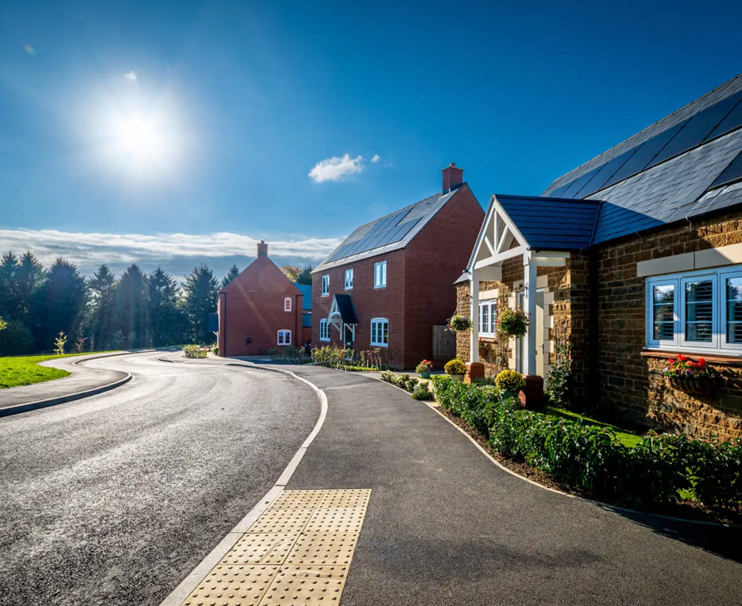 New build red brick homes with solar panels on a sunny residential street surrounded by lush green trees and landscaped gardens.
