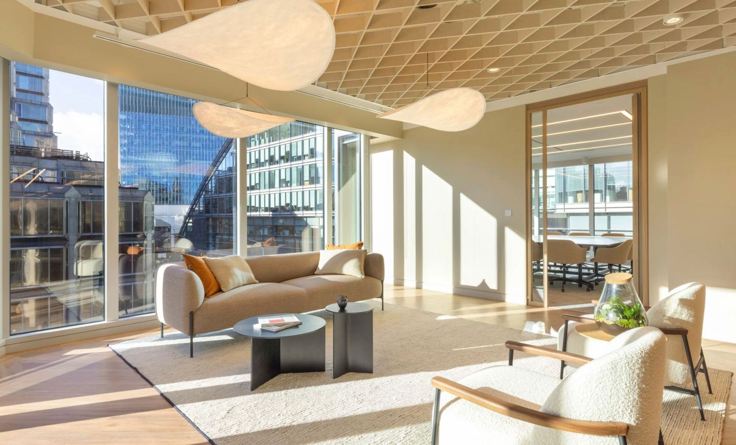 Modern luxury lounge with floor-to-ceiling windows, pendant lights, beige sofa, and wooden accents in a sunlit office space.