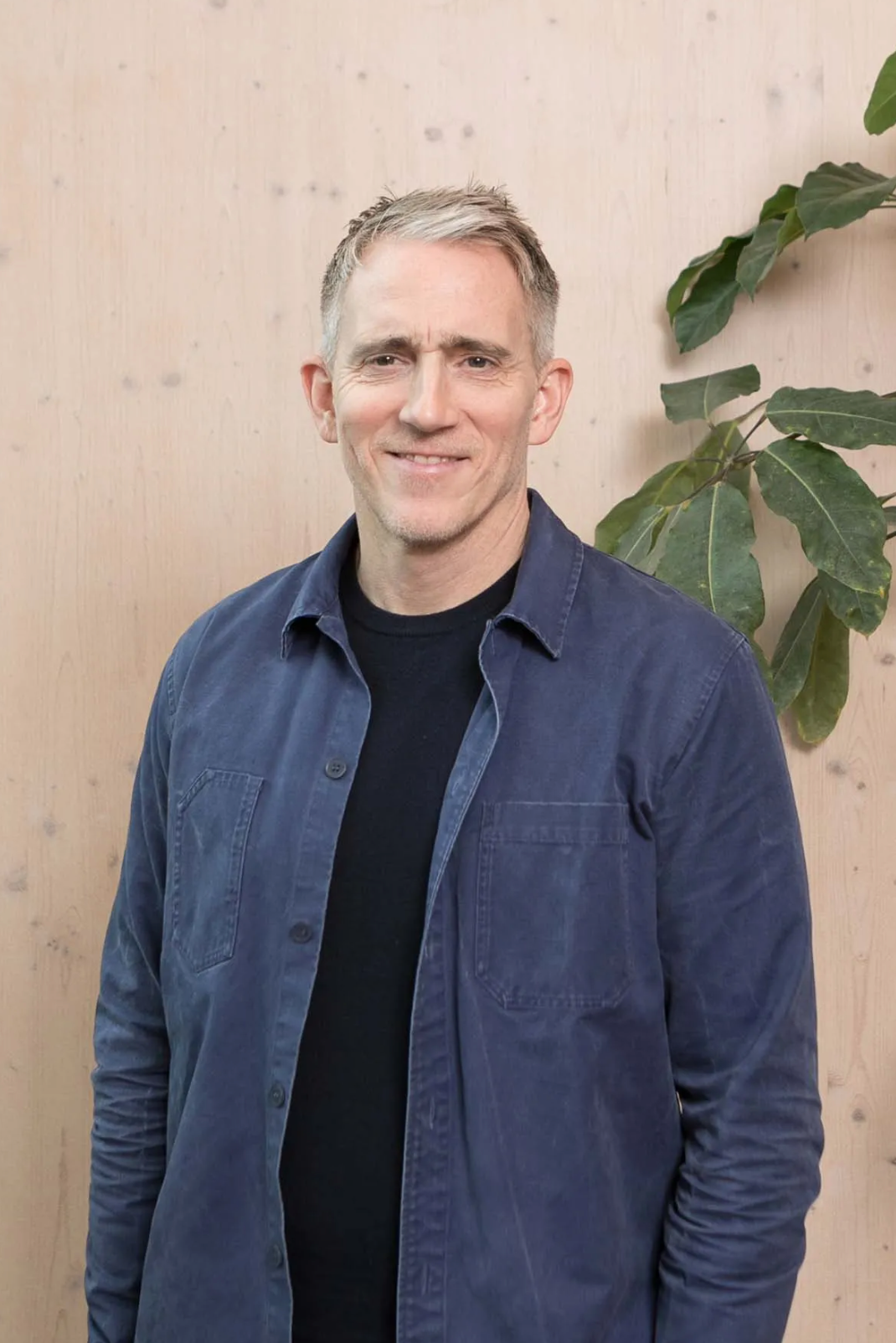 A smiling middle-aged man with gray hair wearing a blue jacket over a black shirt, standing near a plant.