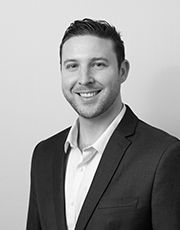 Professional black and white headshot of a smiling young businessman in a suit jacket and collared shirt.