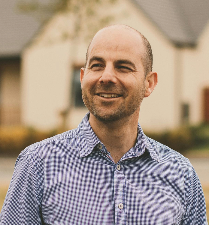 Smiling bald man in a blue checkered shirt standing outdoors near a house on a sunny day.