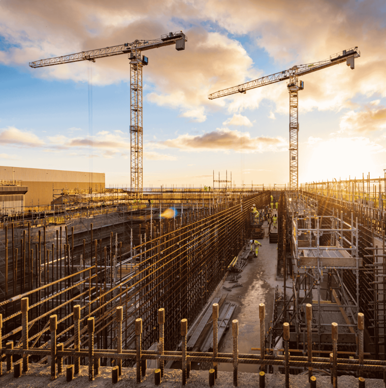 Large construction site with steel rebar framework, scaffolding, and two tower cranes against a dramatic sunset sky.
