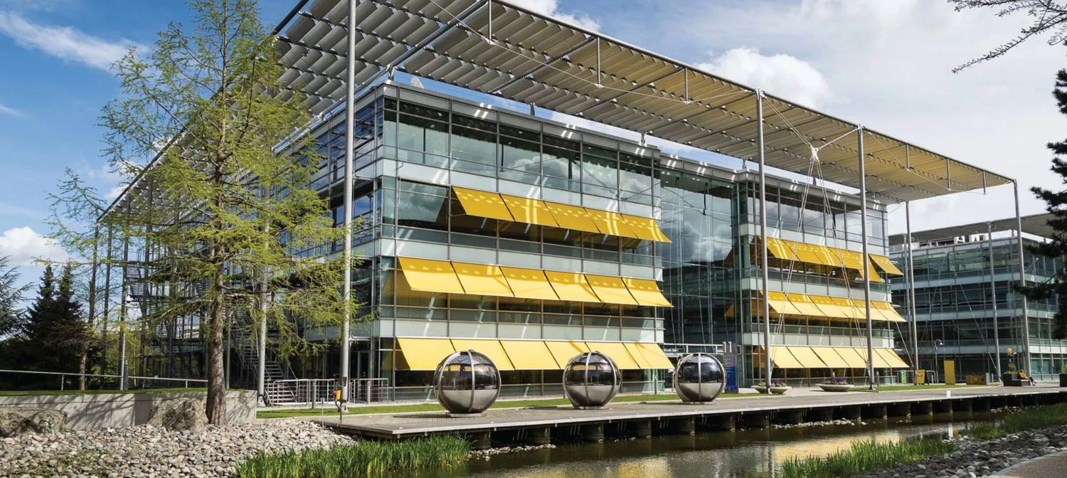 Modern glass office building with yellow awnings reflected in a pond, surrounded by trees on a sunny day.