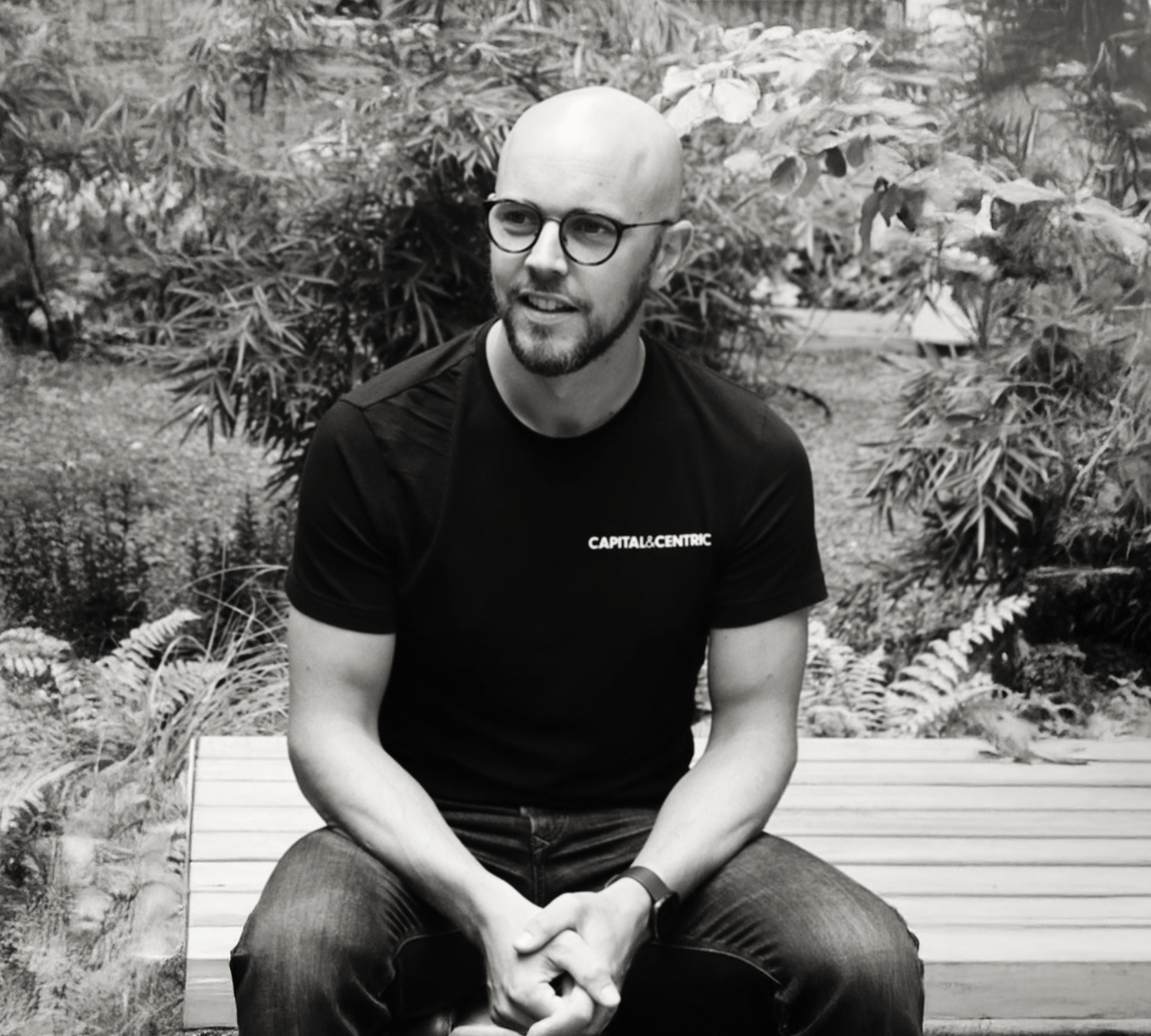 Bald man with glasses wearing a Capital & Centric t-shirt sits on a bench surrounded by lush greenery.
