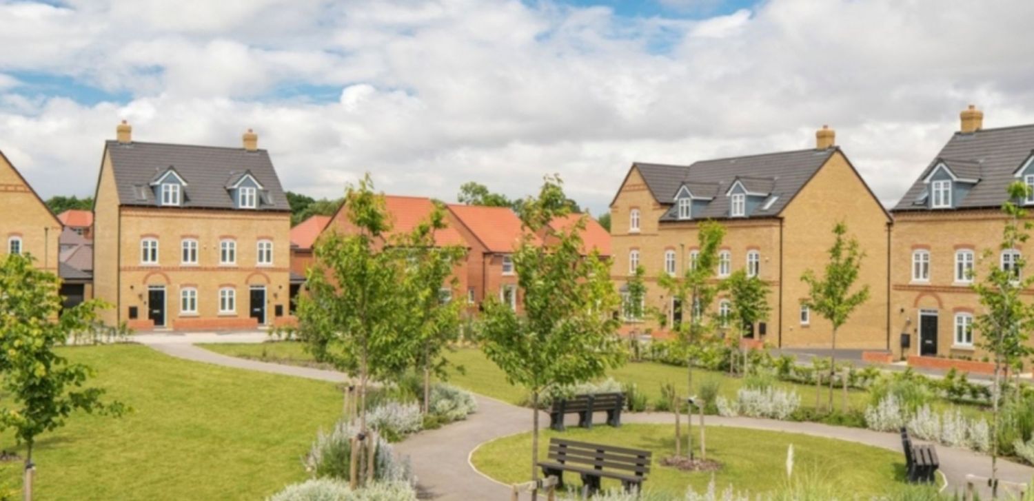 Modern residential housing development with green communal garden, benches, and landscaped pathways under a partly cloudy sky.