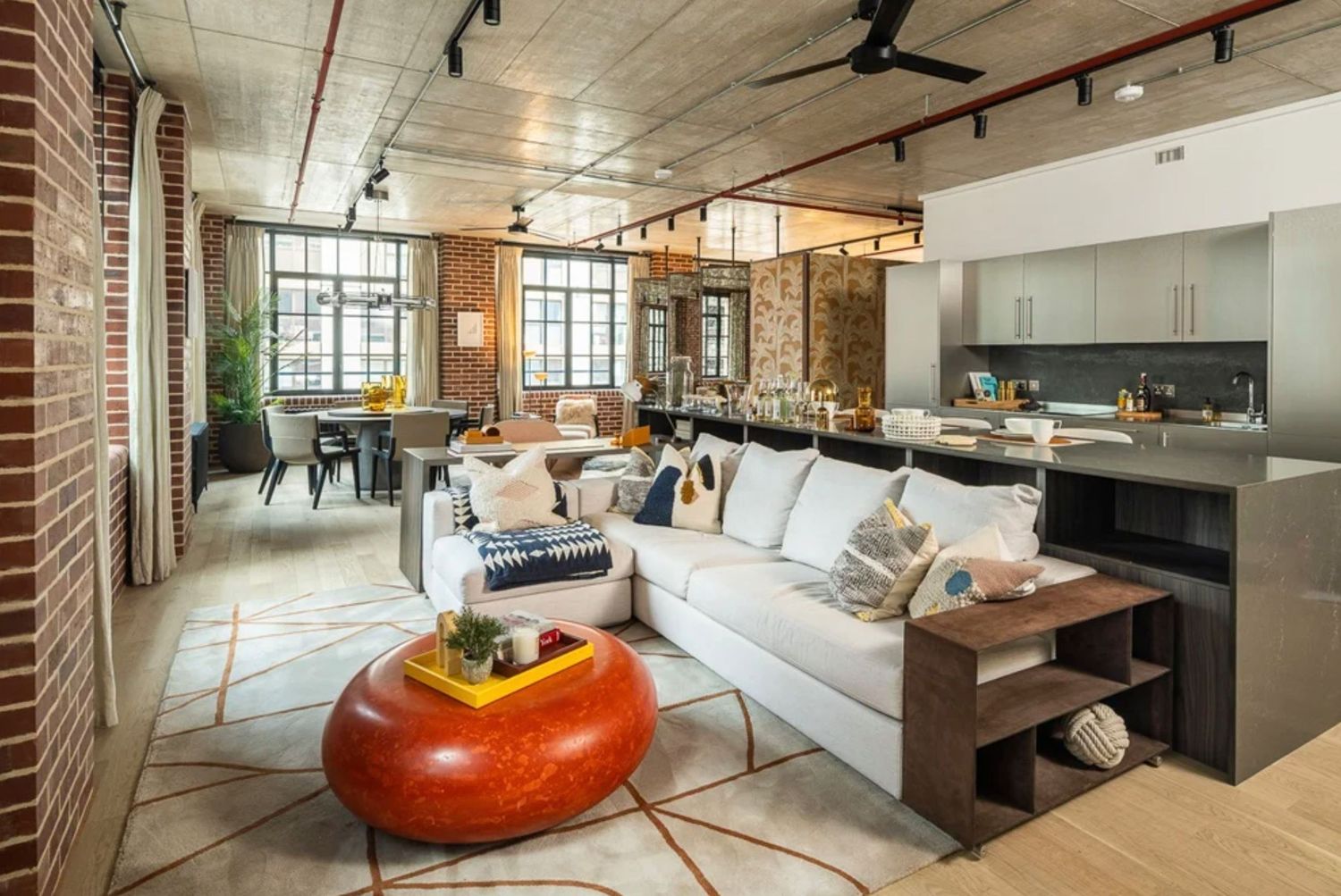 Modern loft living room with white sectional sofa, red ottoman, brick walls, open kitchen, and industrial ceiling in an urban apartment.