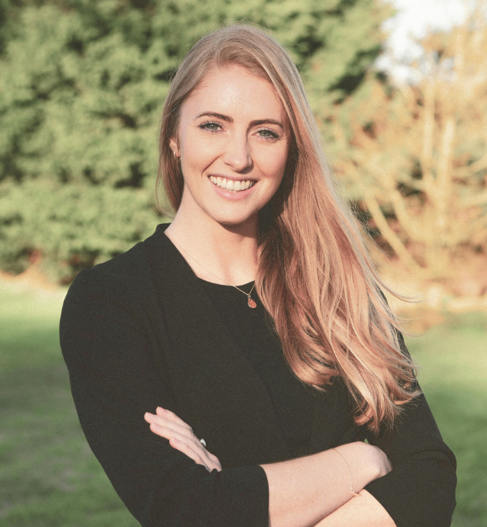 Young woman with long blonde hair smiling outdoors, wearing a black top with arms crossed in a park setting.