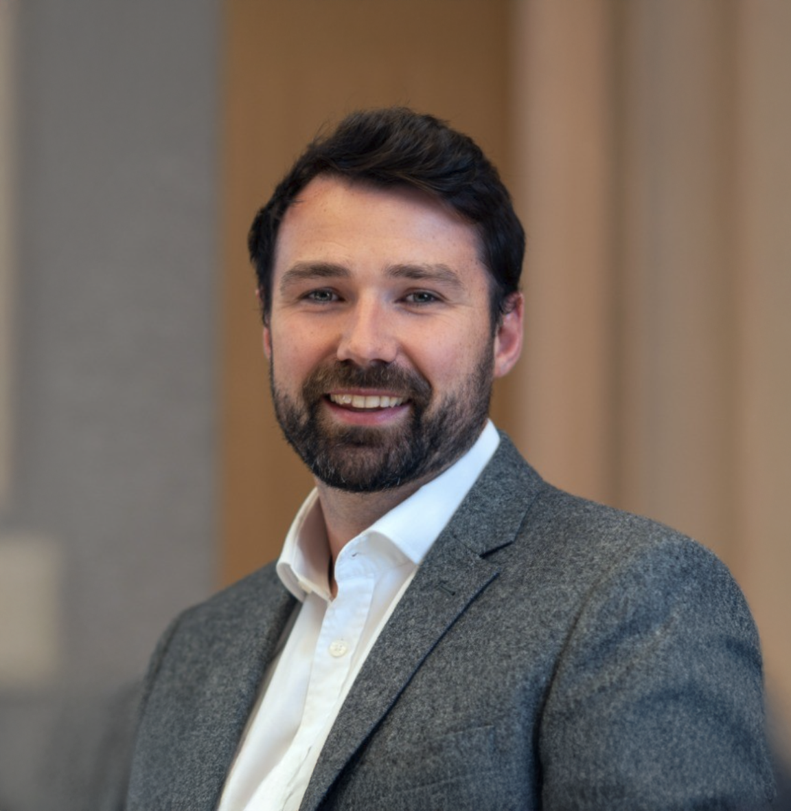 Professional man with beard smiling in gray blazer and white shirt, posed for a corporate headshot photo.