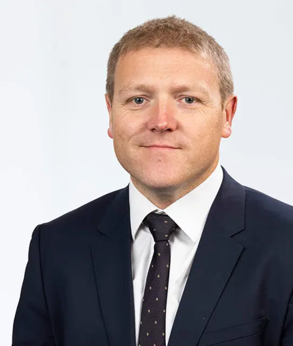 Professional headshot of a middle-aged man in a dark suit and tie against a light gray background.