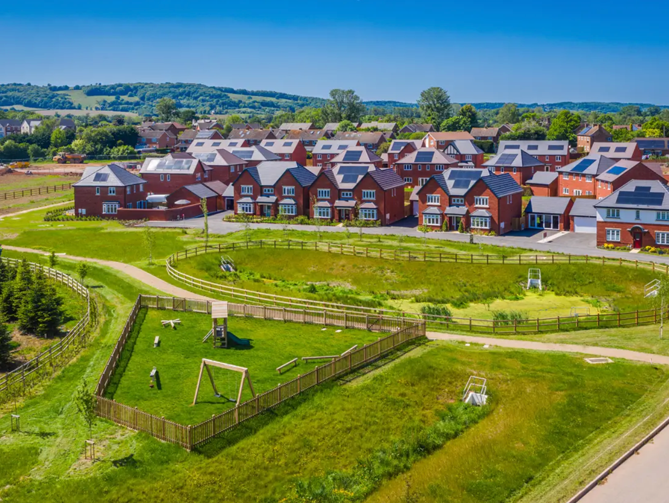 Aerial view of modern red brick housing development with solar panels, green lawns, and fenced grounds on a sunny day.