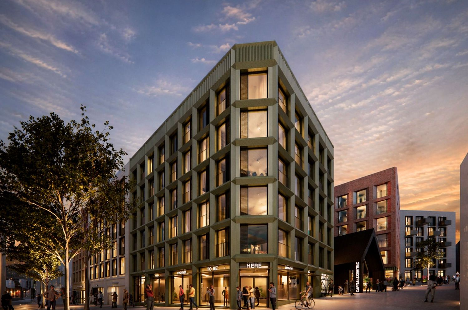 Modern green-facade mixed-use building at dusk with ground-floor retail, large windows, and urban streetscape in warm evening light.