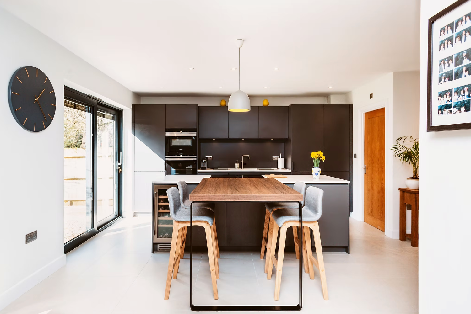 Modern open concept kitchen and dining area with black cabinets, wooden table and white floors in minimalist style.