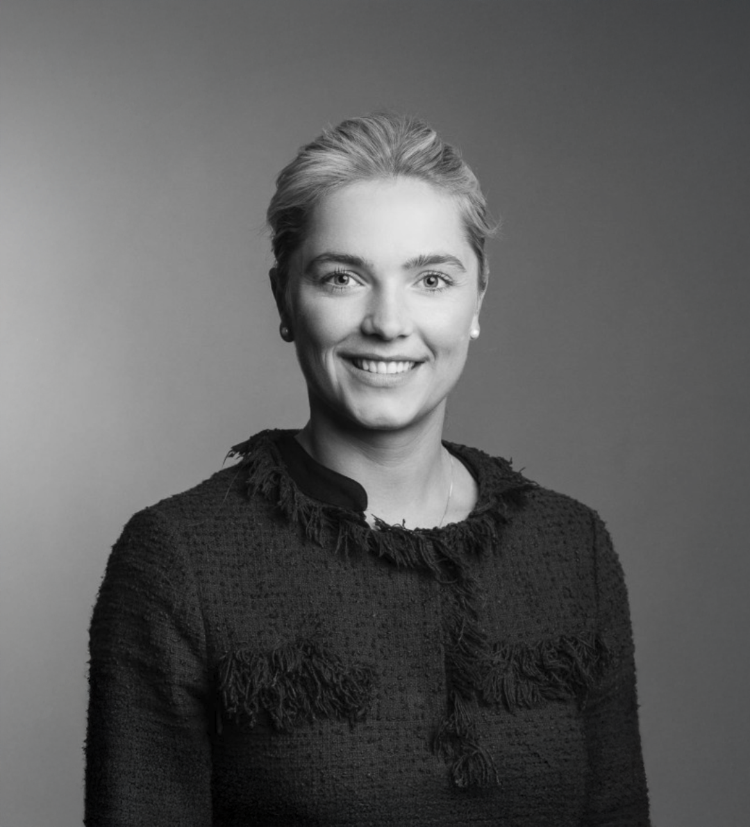Black and white professional headshot of a smiling blonde woman in a textured dark blazer against a neutral background.
