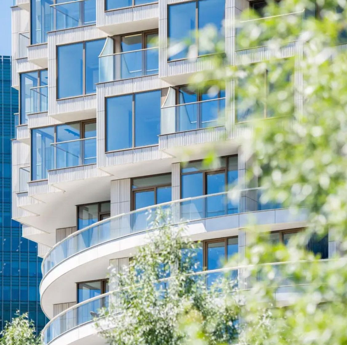 Modern high-rise apartment building with curved glass balconies and blue reflective windows surrounded by lush green trees.