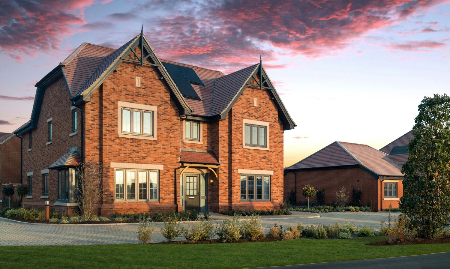 Large red brick detached house with dark roof, bay windows, and manicured landscaping at dusk.