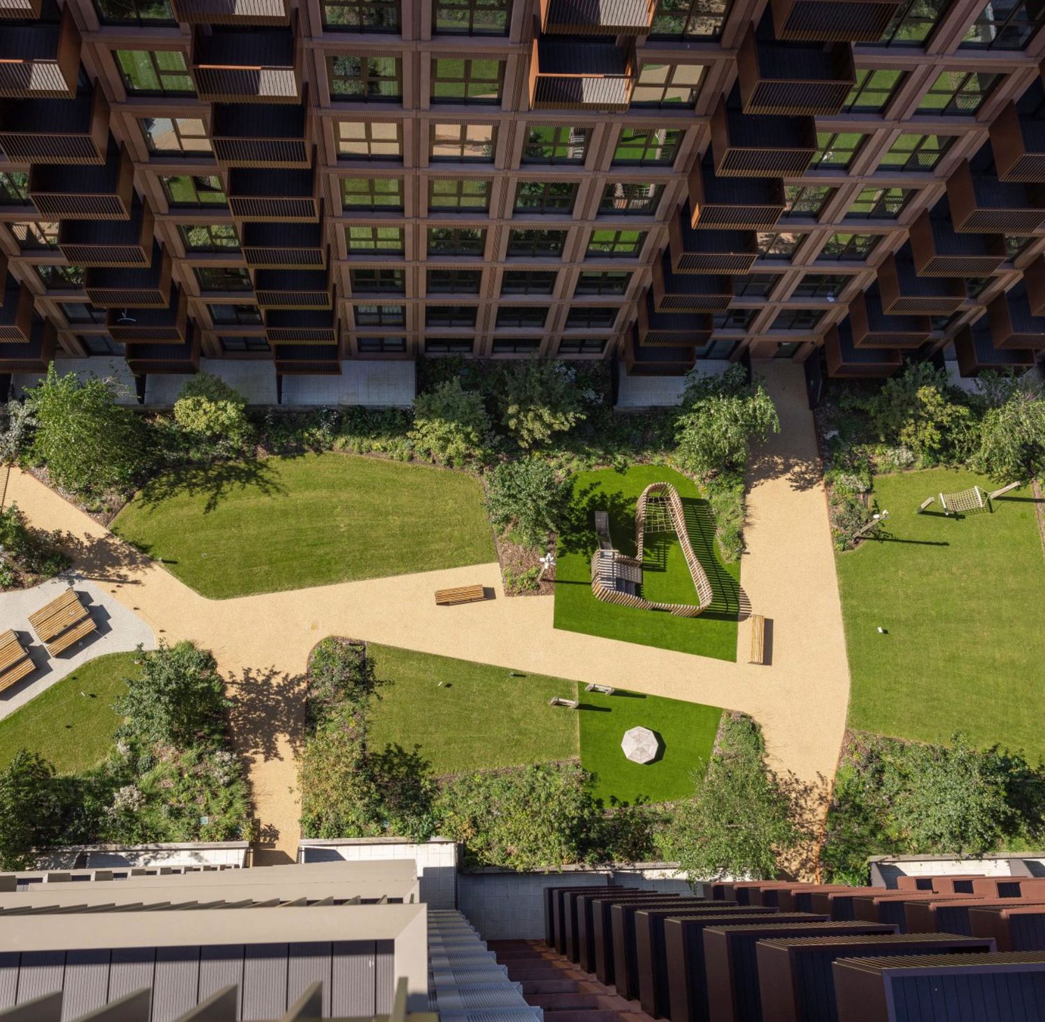 Aerial view of a modern residential courtyard with lush green gardens, pathways, and wooden benches surrounded by a grid-facade building.