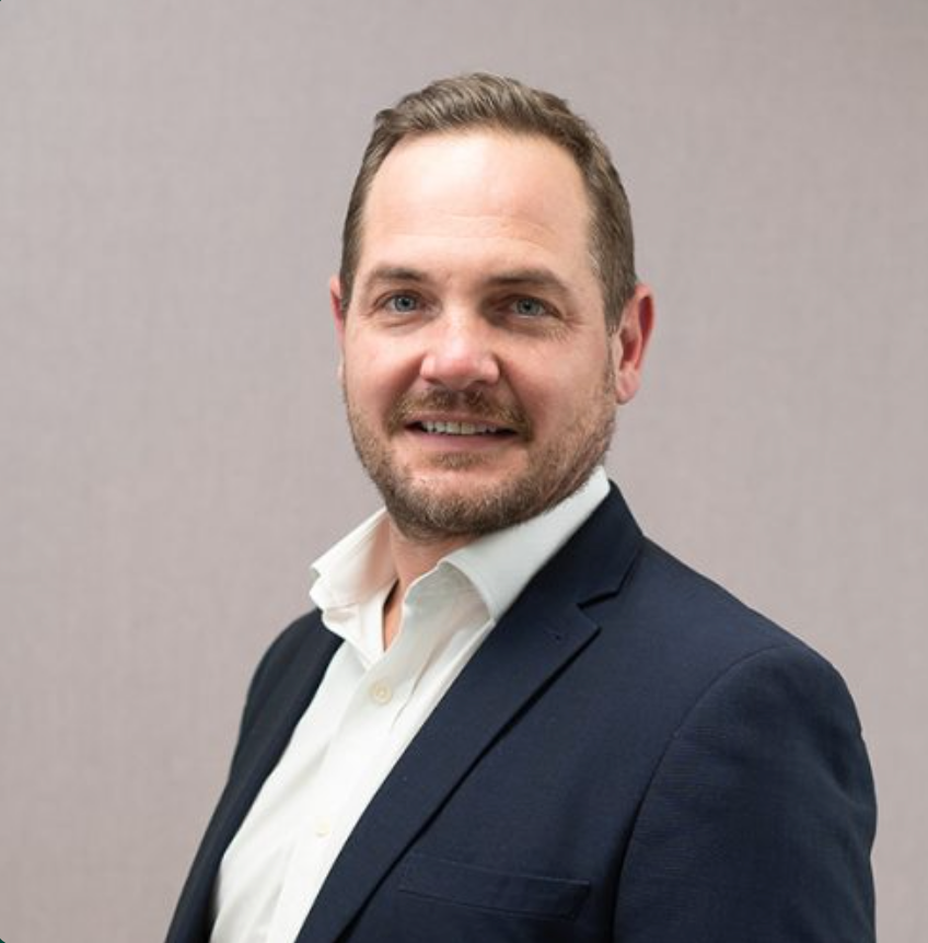 Professional businessman in navy blazer and white shirt smiling confidently against a neutral gray background.