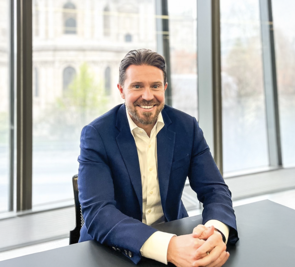 Smiling businessman in navy blazer sits at desk with clasped hands, modern office with historic building view.
