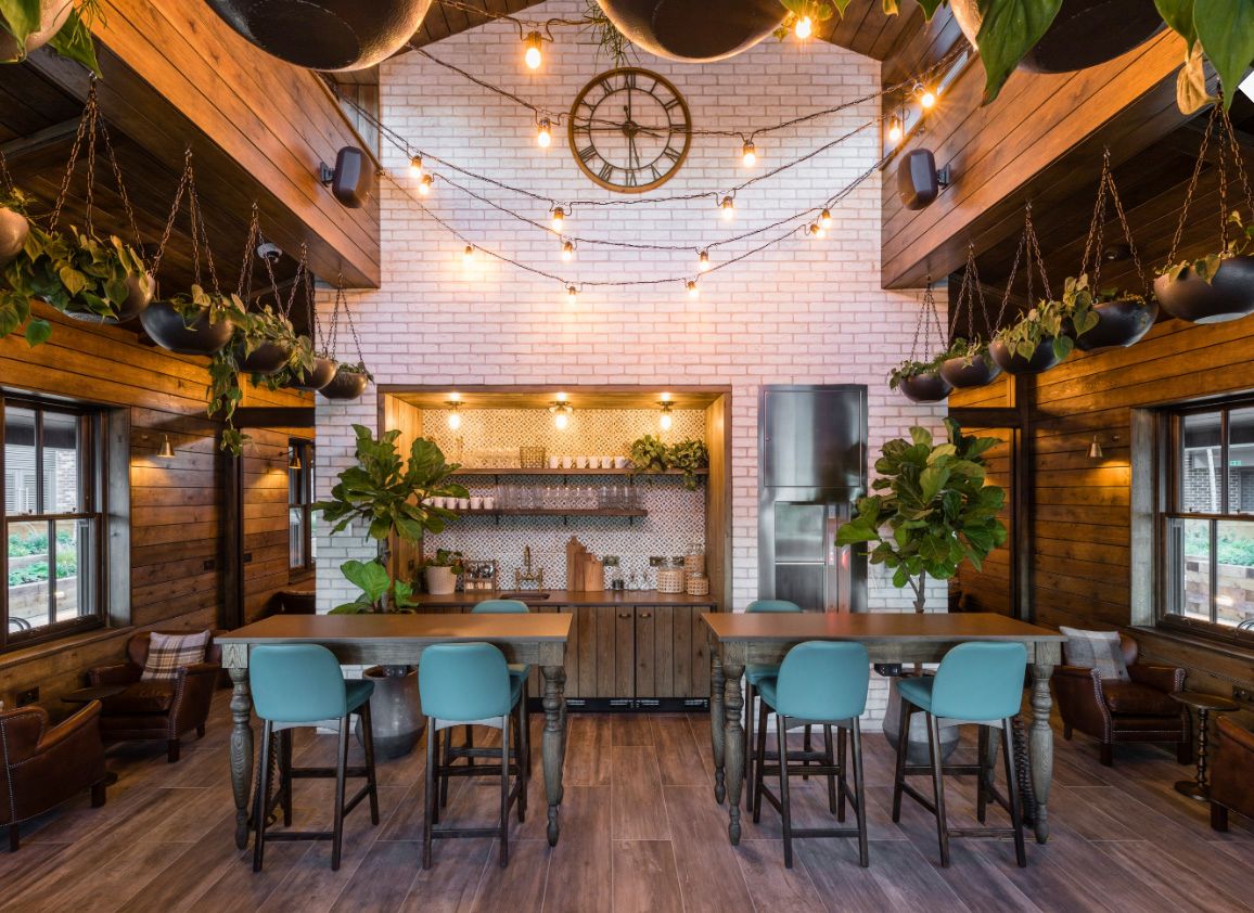 Rustic restaurant interior with teal bar stools, string lights, hanging plants, and white brick wall backdrop.