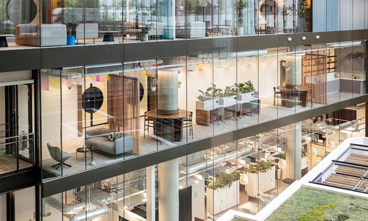 Modern multi-story office building interior with glass walls, green plants, and stylish open-plan workspaces.