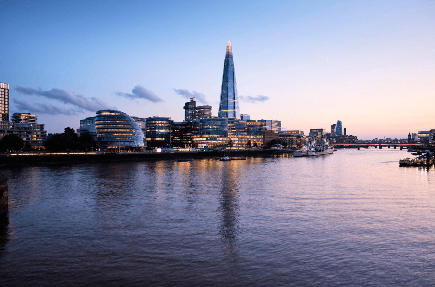 London skyline at dusk featuring The Shard skyscraper reflecting on the River Thames with city lights glowing.