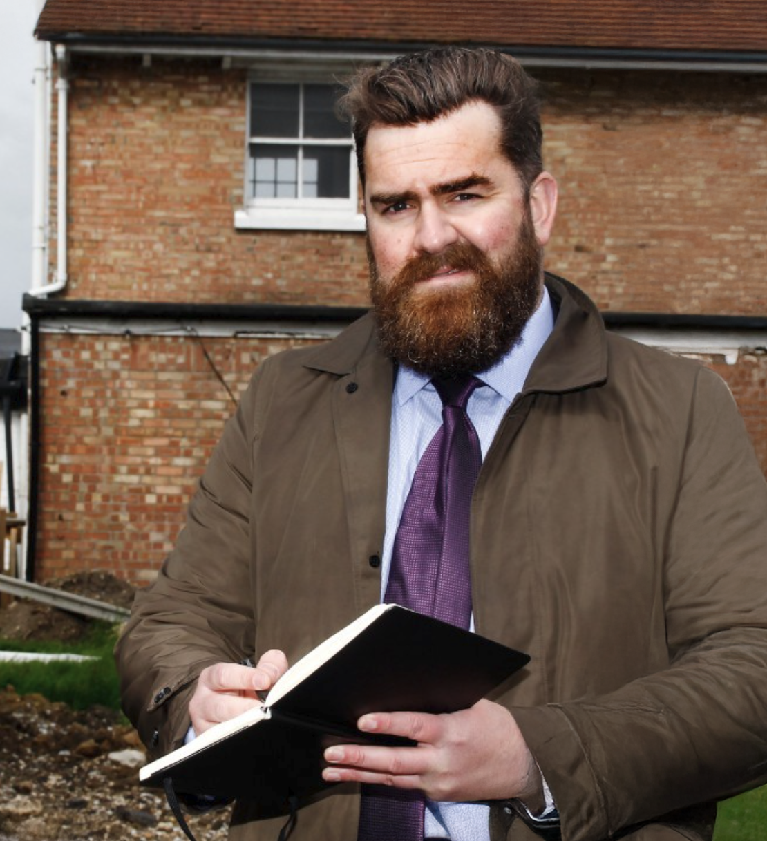 Bearded man in brown jacket and purple tie writing in notebook outdoors near brick building.