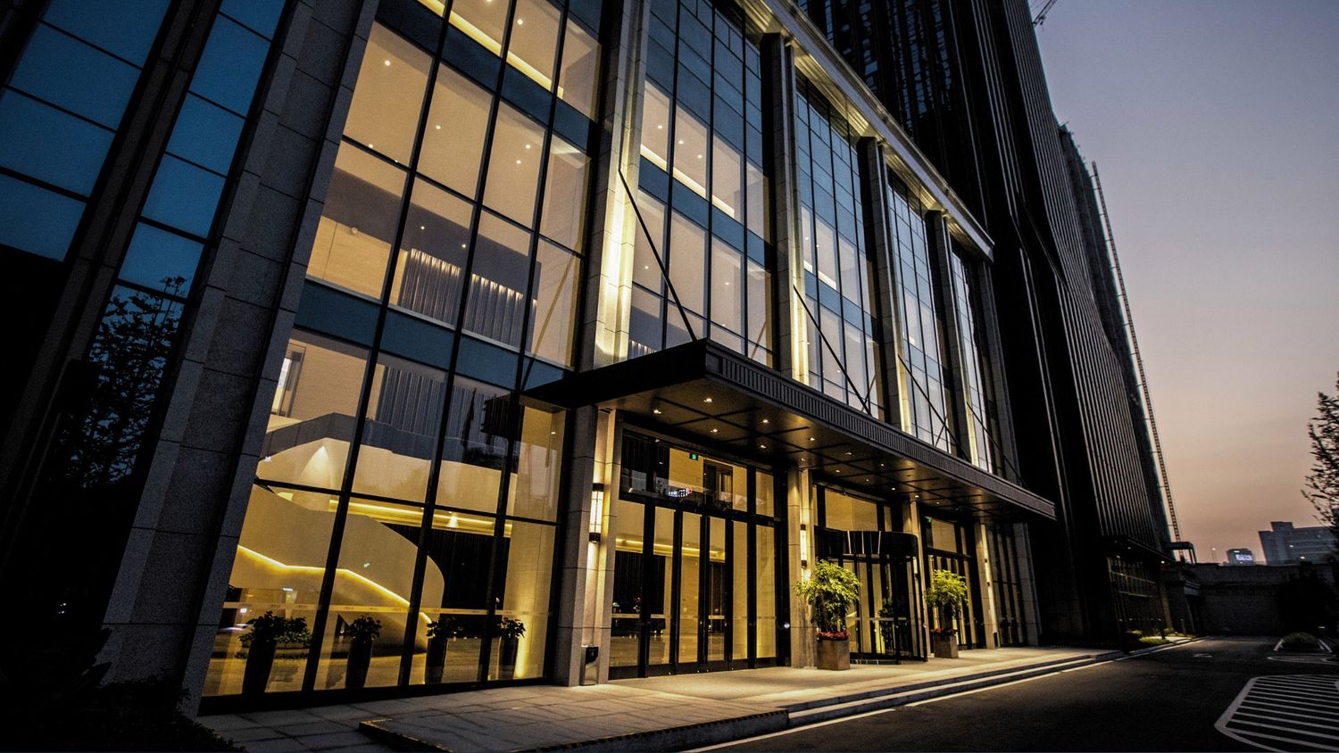 Modern glass office building exterior illuminated at dusk with elegant entrance and landscaping.