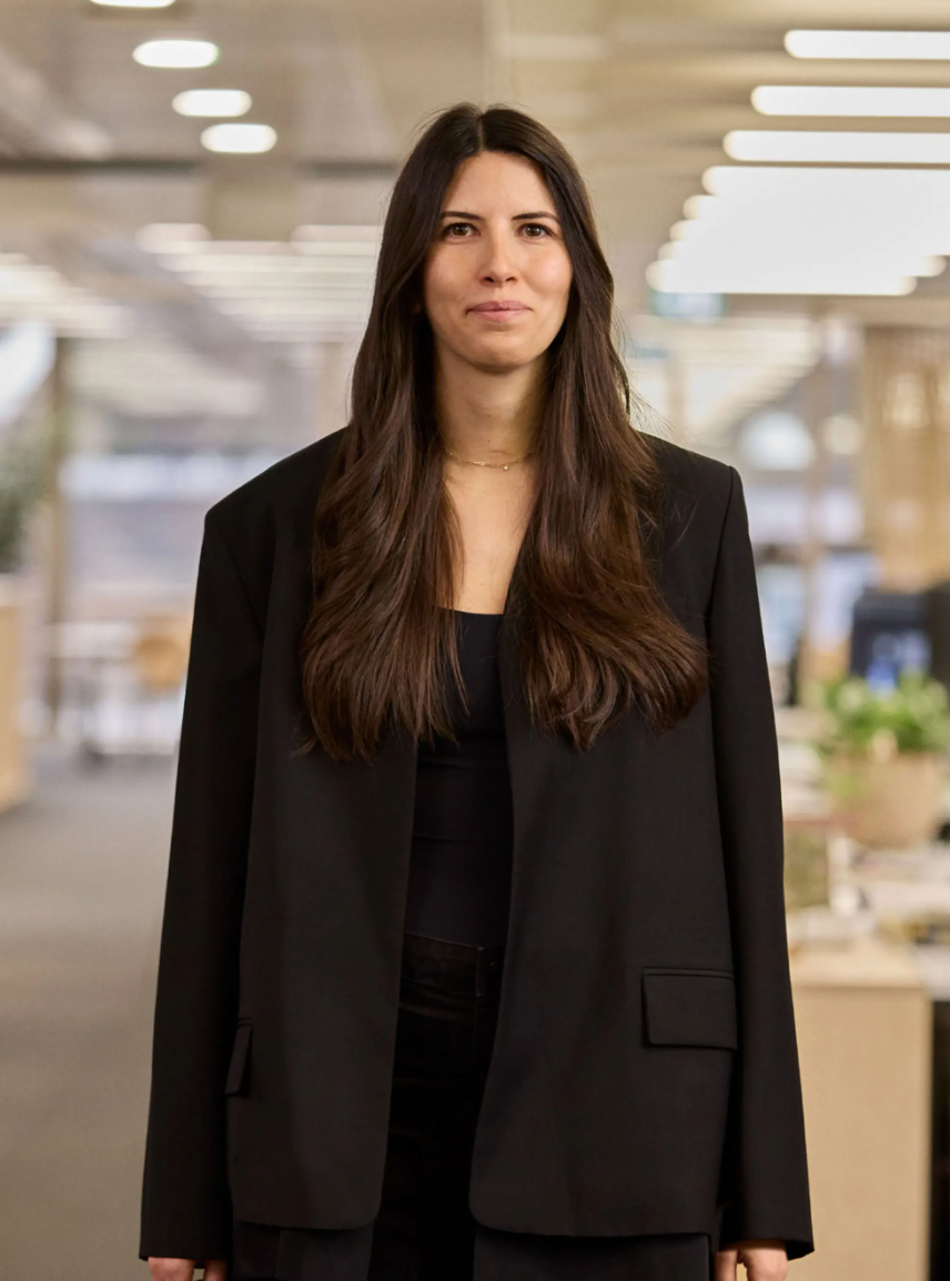 A professional woman in a black blazer stands confidently in a modern office environment with long dark hair.
